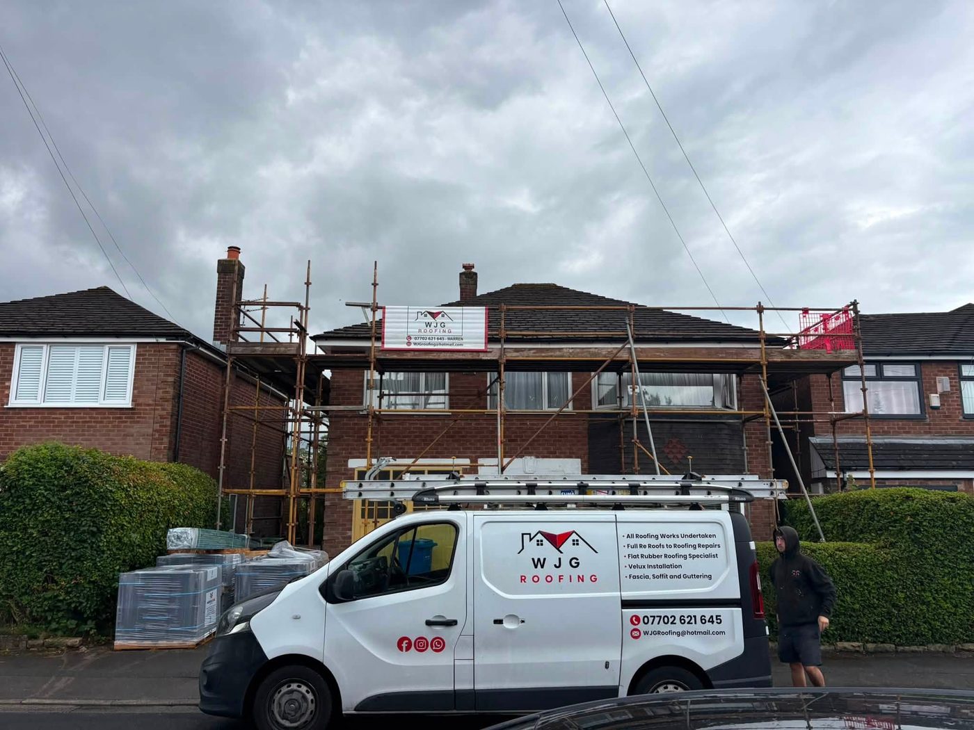 WJG Roofing van in full livery parked outside a scaffolded semi-detached house mid re-roof, with a branded WJG Roofing banner fixed to the scaffold and pallets of new tiles waiting on the drive