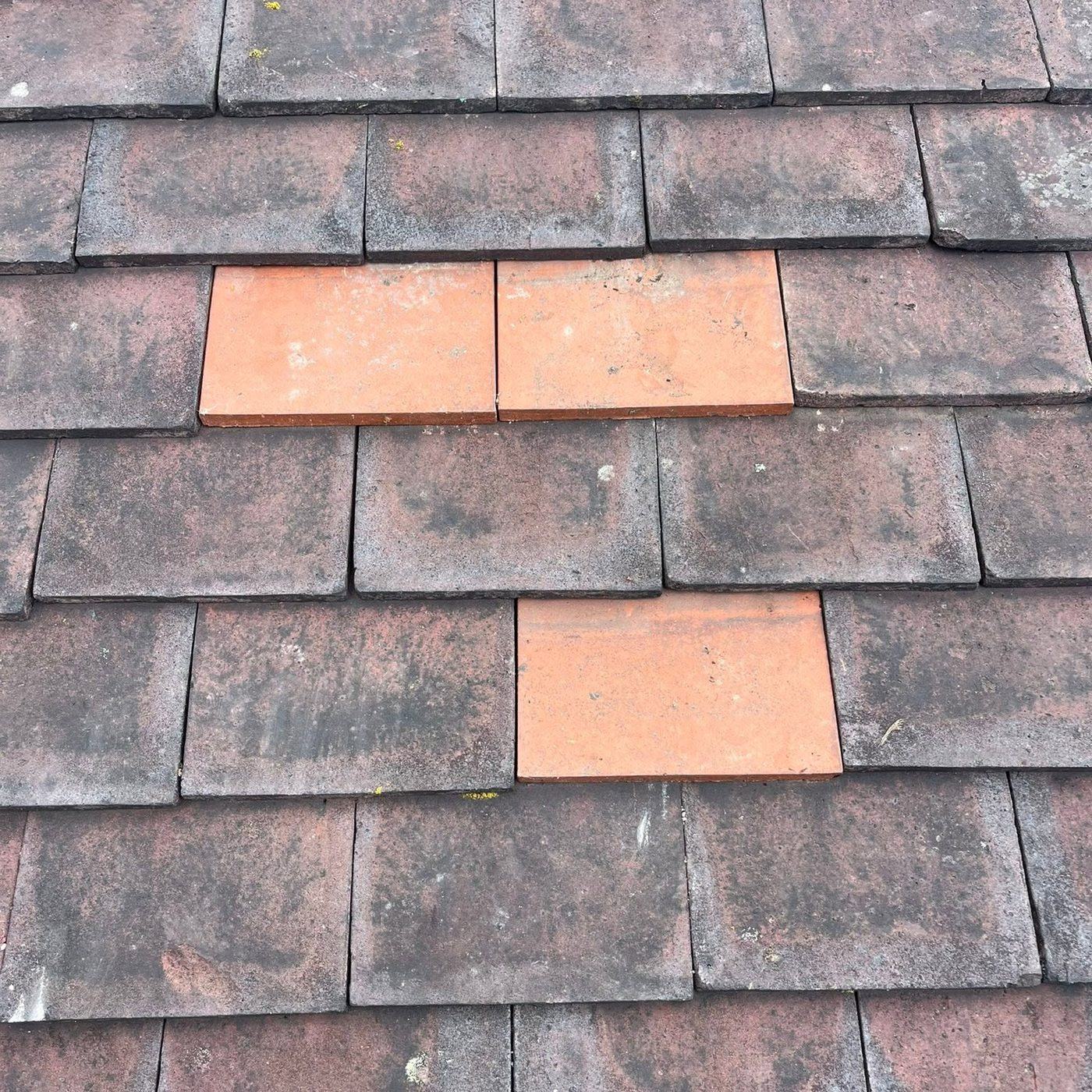 Close up of a plain tiled roof with three fresh terracotta clay tiles slotted in among the weathered grey and red tiles, two sitting side by side in one course and a third in the course below