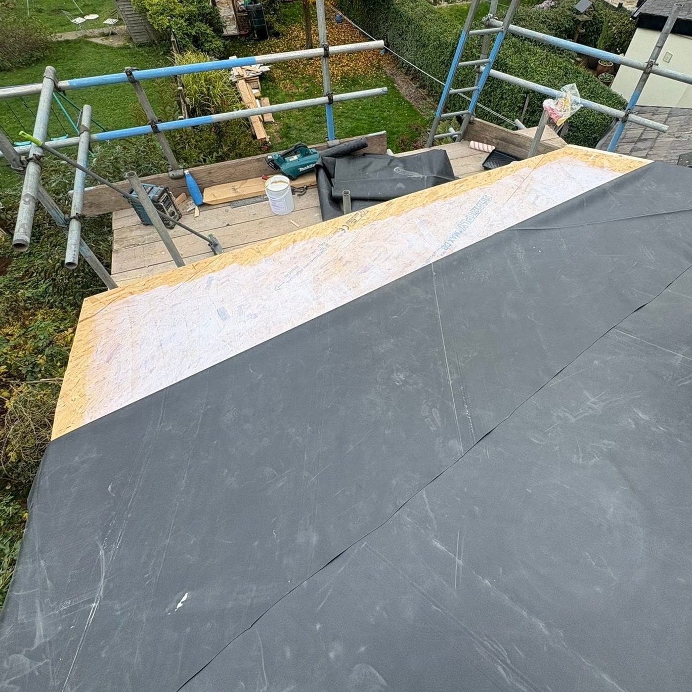 Large single sheet EPDM rubber membrane being laid over a new timber deck on a rear extension roof, with scaffolding around the edge